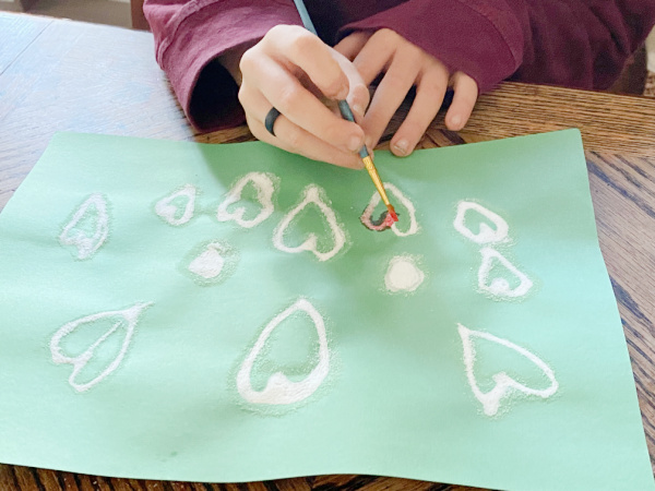 Anak melukis lem dan garam hati di atas kertas konstruksi hijau untuk membuat kegiatan seni garam dan cat air Valentines.