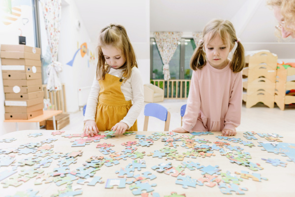 Two young preschool girls are putting together a large puzzle. There are puzzle pieces spread out on a table in front of them. These 6 DIY puzzles for kids to make and do will help brain development and learning.