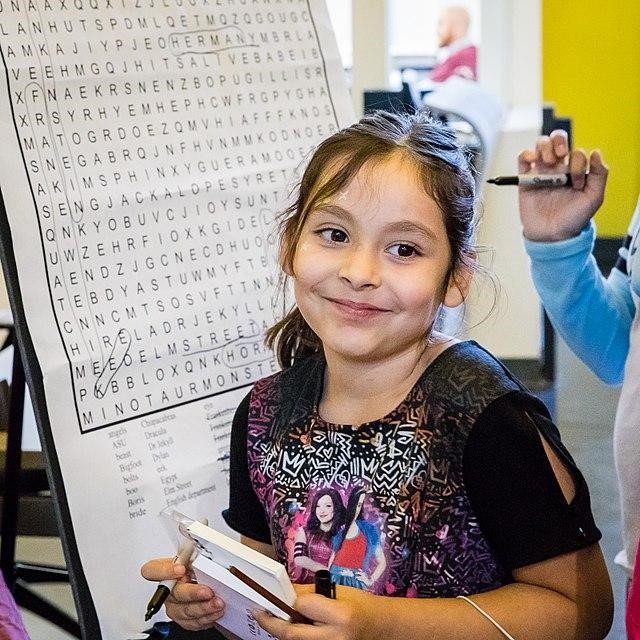 Girl smiling with large classroom word search poster behind her. Article is about top 5 benefits of word searches for your kids.
