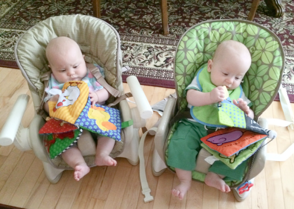 Two babies reading books in child seats. Article details the reality of life with twins for twin moms.