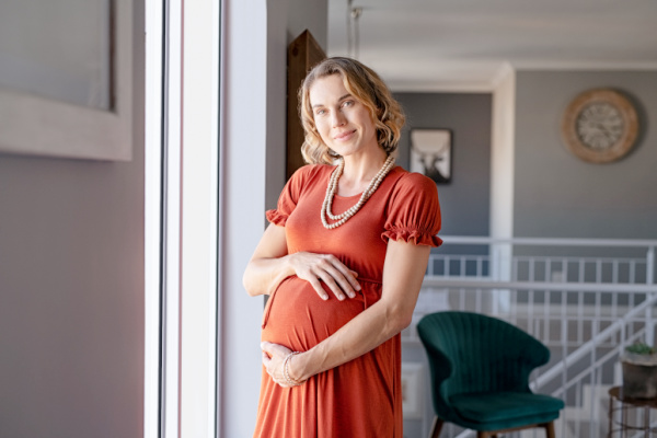Pregnant woman is smiling and holding her belly while standing in front of a window. 