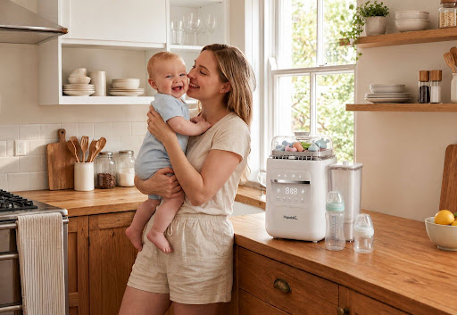 Happy mother holding smiling baby beside Papablic SafeguardPlus™ Baby Bottle Washer System (P8Plus) and baby bottles on kitchen counter